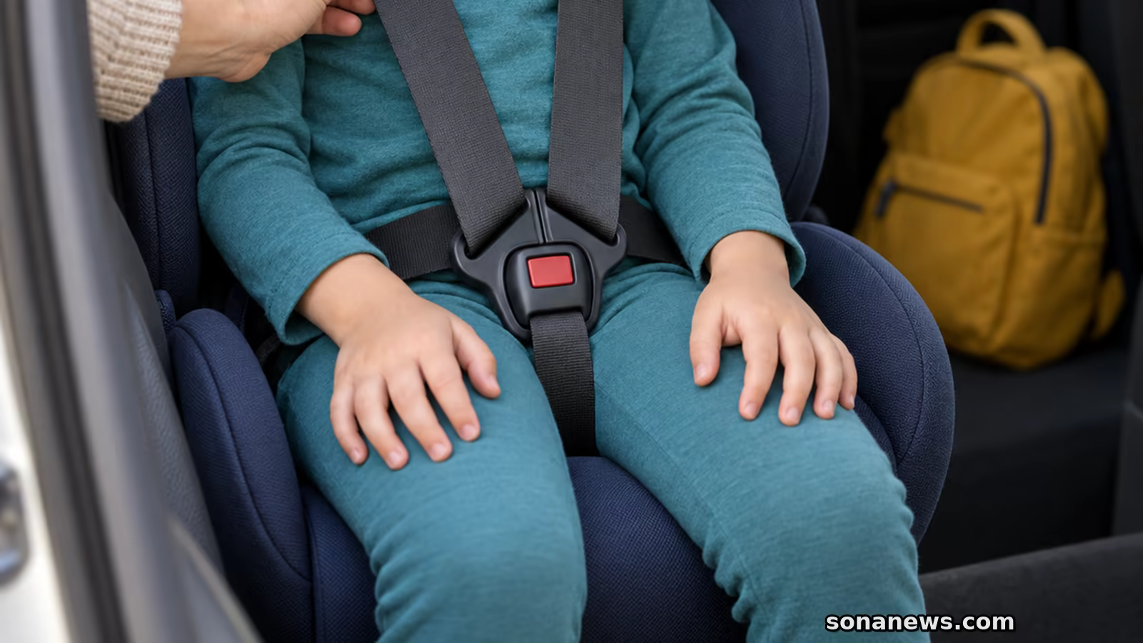 Mão adulta verifica o arnês de uma cadeirinha antes do trajeto escolar, com mochila amarela no piso traseiro.